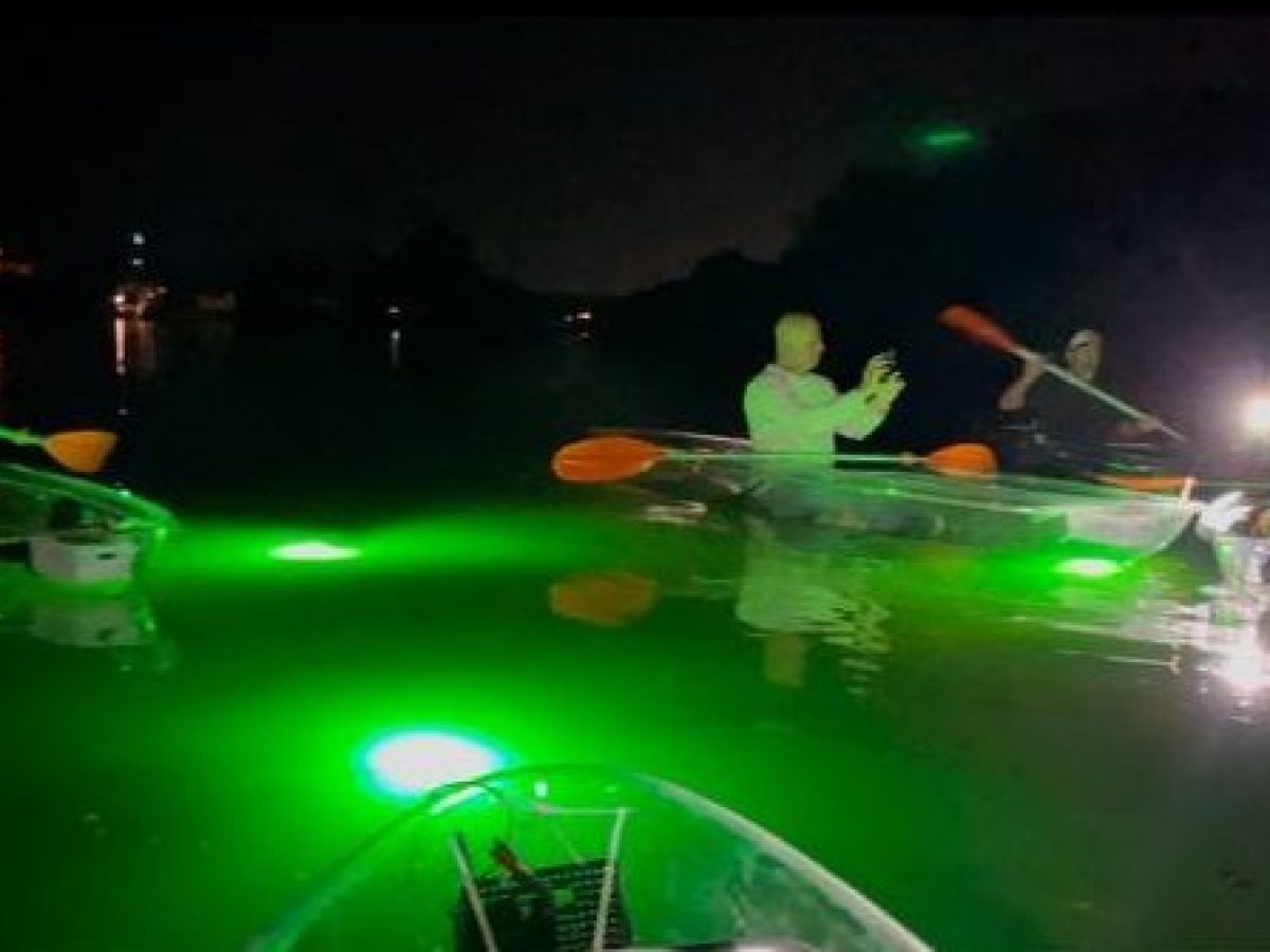 Group of kayakers paddling in glass-bottom kayaks with green underwater lights at night, creating a glowing effect on the calm water.