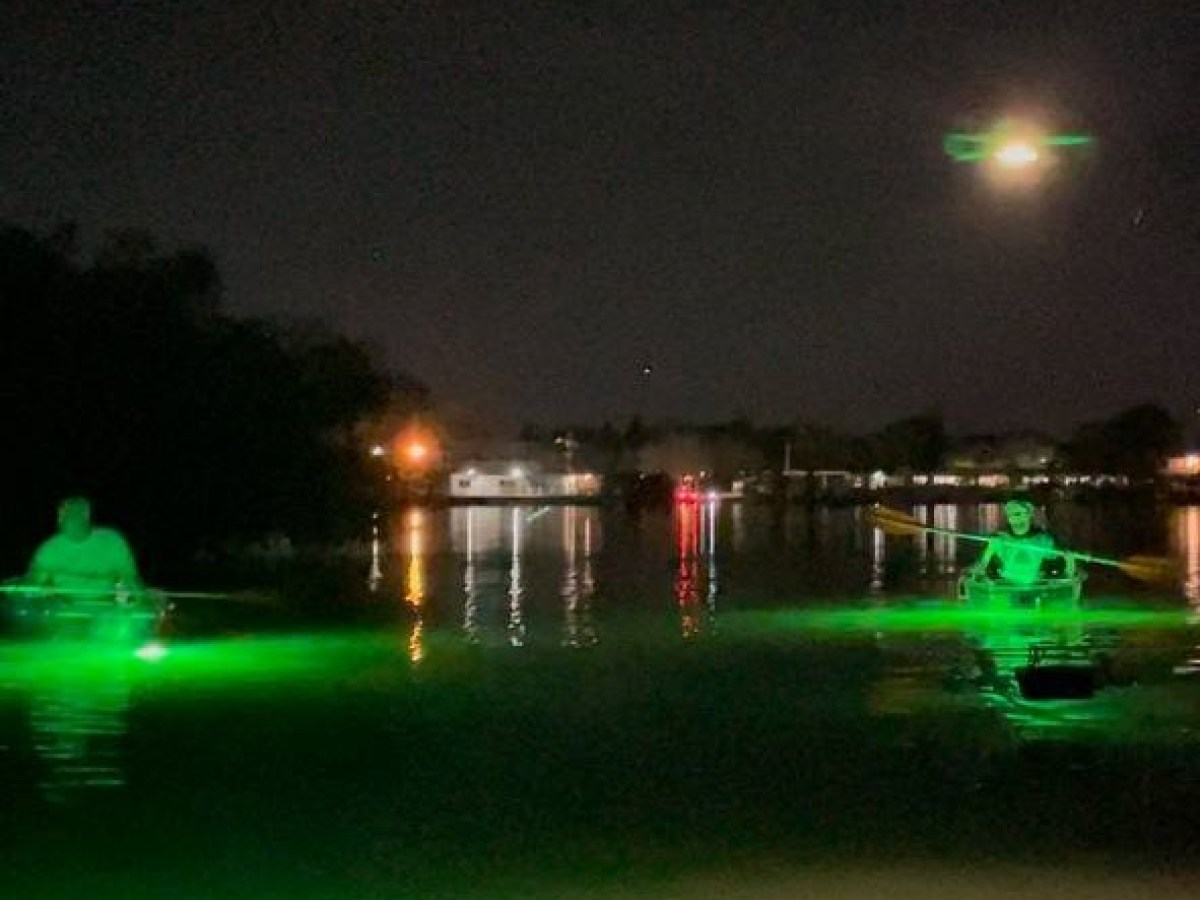 Two kayakers paddling illuminated glass-bottom kayaks with green lights under a glowing full moon, surrounded by calm waters and distant waterfront lights.