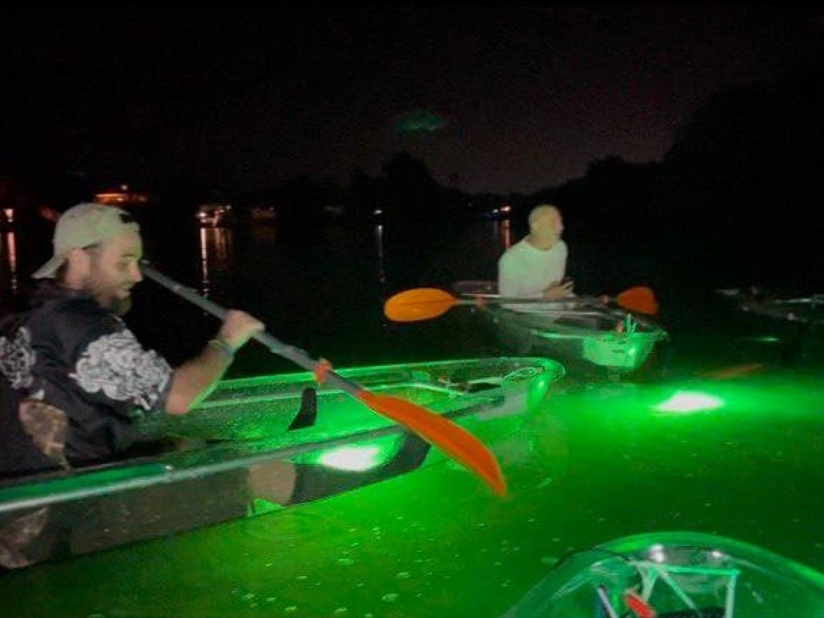 Two individuals kayaking in illuminated glass-bottom kayaks with green underwater lights at night, surrounded by calm waters.