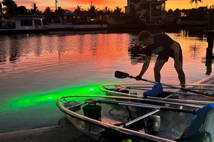 Person kneeling by paddle boards at sunset, waterfront houses in view.