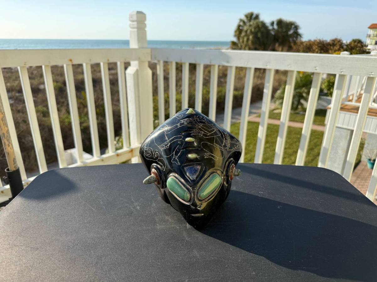 Black ceramic item with carvings on a table, sea view and greenery in the background.