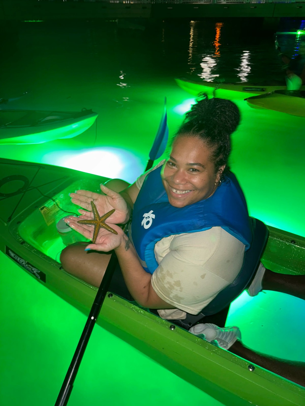 Woman in kayak at night holding a starfish, surrounded by glowing green water.