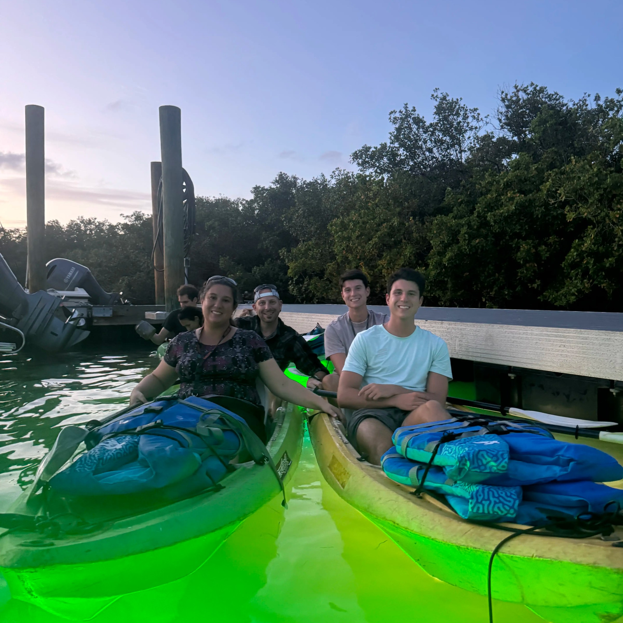Four people in glowing kayaks on water near a dock at dusk.