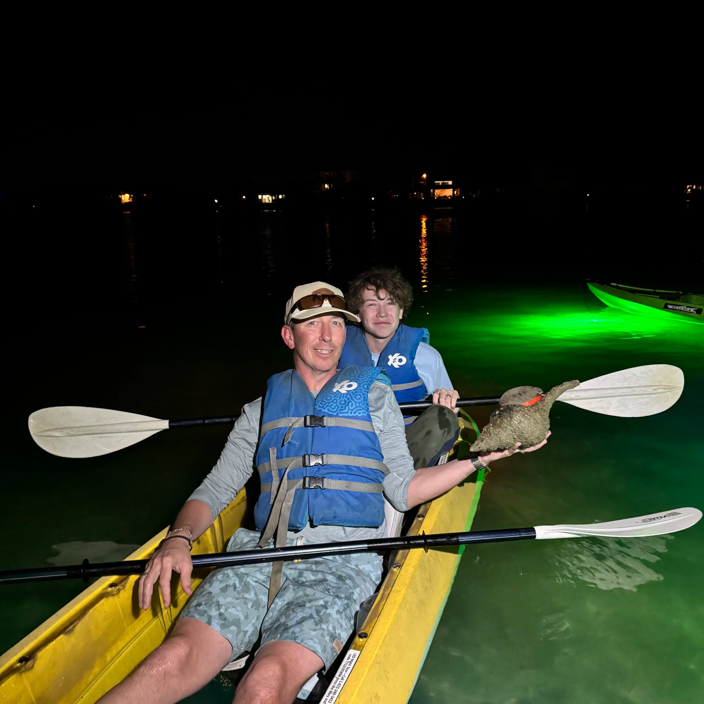 Two people in a kayak at night, wearing life vests, illuminated by green light.