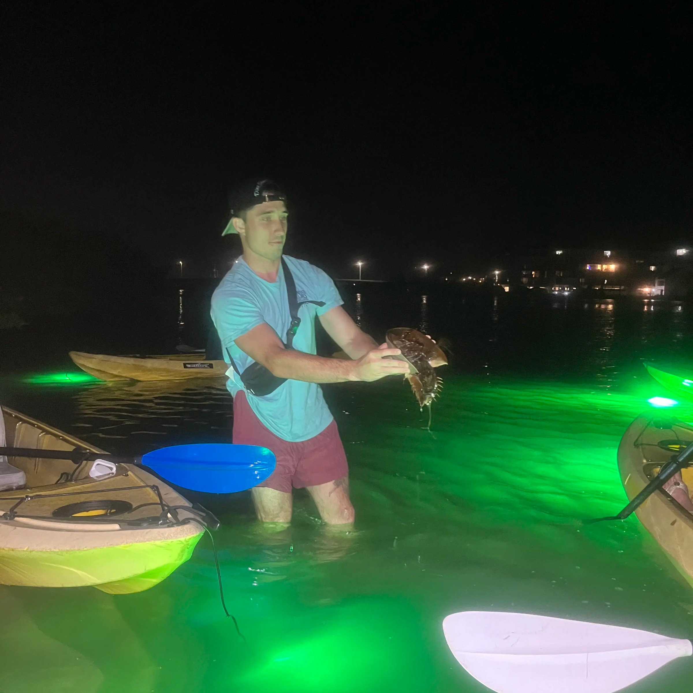 Person holds a fish in knee-deep water near kayaks, under green lights at night.